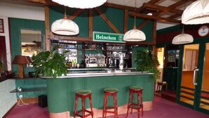 Zona de bar con un letrero verde que dice "Heineken", tres taburetes de madera y lámparas colgantes.
