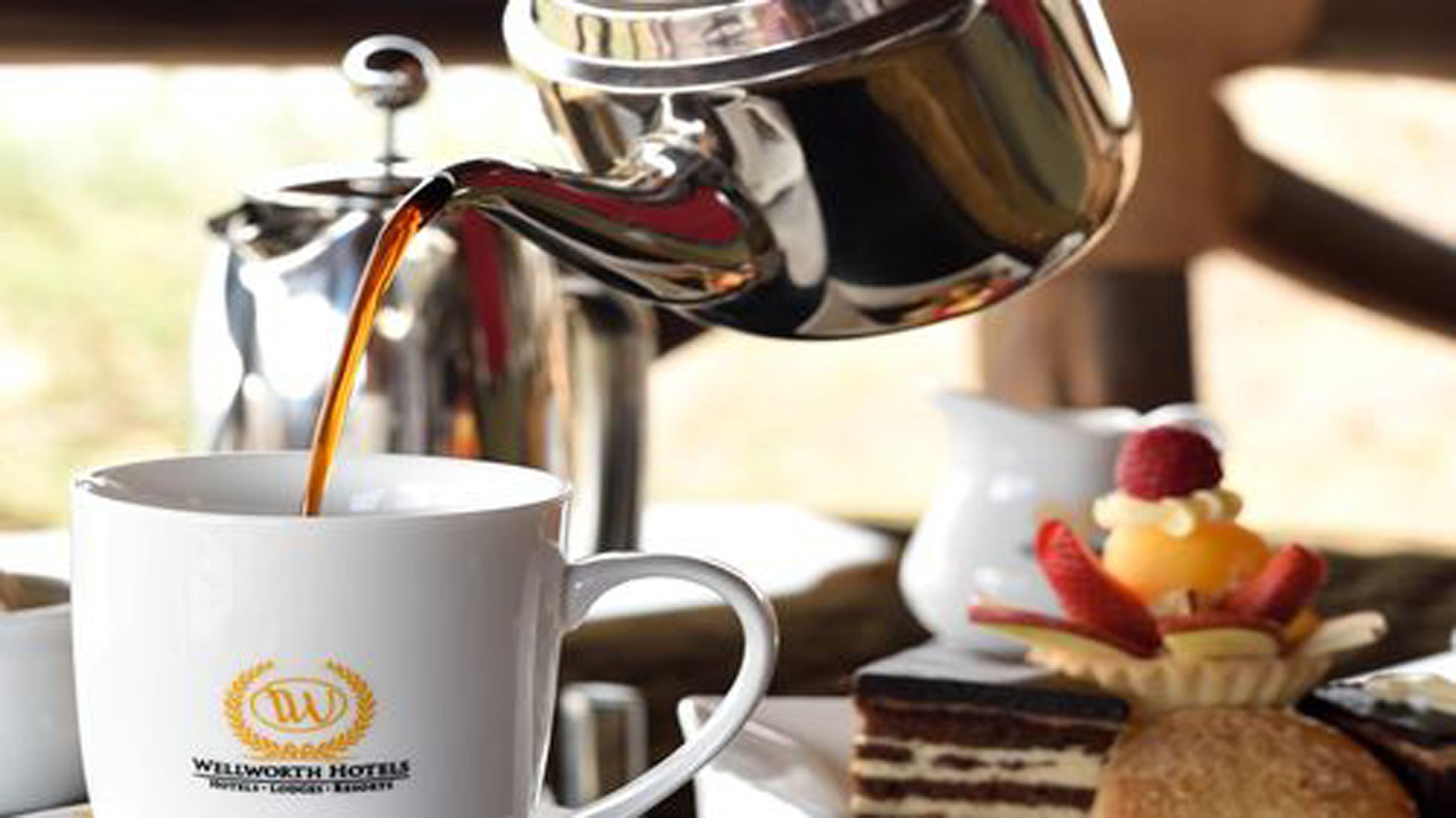 A coffee pot pouring into a mug with a hotel logo, a dessert plate, and a cake on a table.