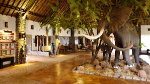 A reception area with a stone pillar, wooden bench, and a statue. There is a large elephant statue with tusks. - Lake Manyara Kilimamoja Lodge (Lake Manyara National Park)