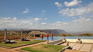 A pool area with lounge chairs, a thatched-roof building, and a mountainous backdrop. - Lake Manyara Kilimamoja Lodge (Lake Manyara National Park)