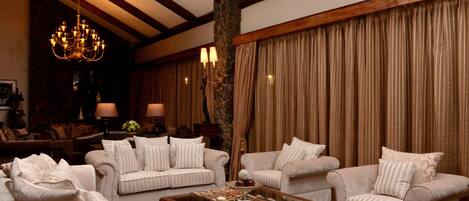 A cozy living room with beige sofas, a coffee table with decorative items, a chandelier, and a large window with curtains.