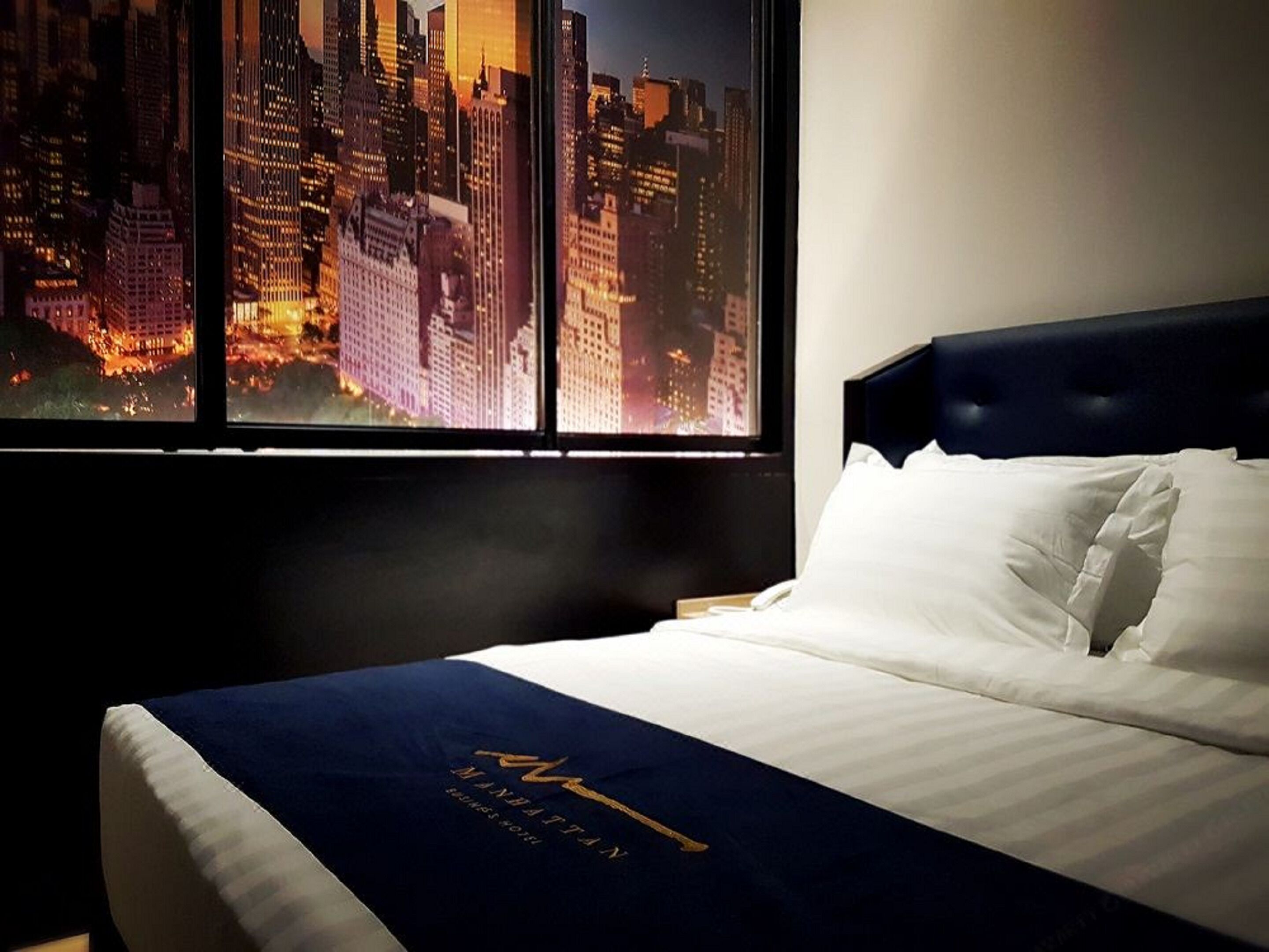 Habitación de hotel con un amplio ventanal que ofrece vistas a la ciudad. La cama tiene un cabecero oscuro y ropa de cama blanca.
