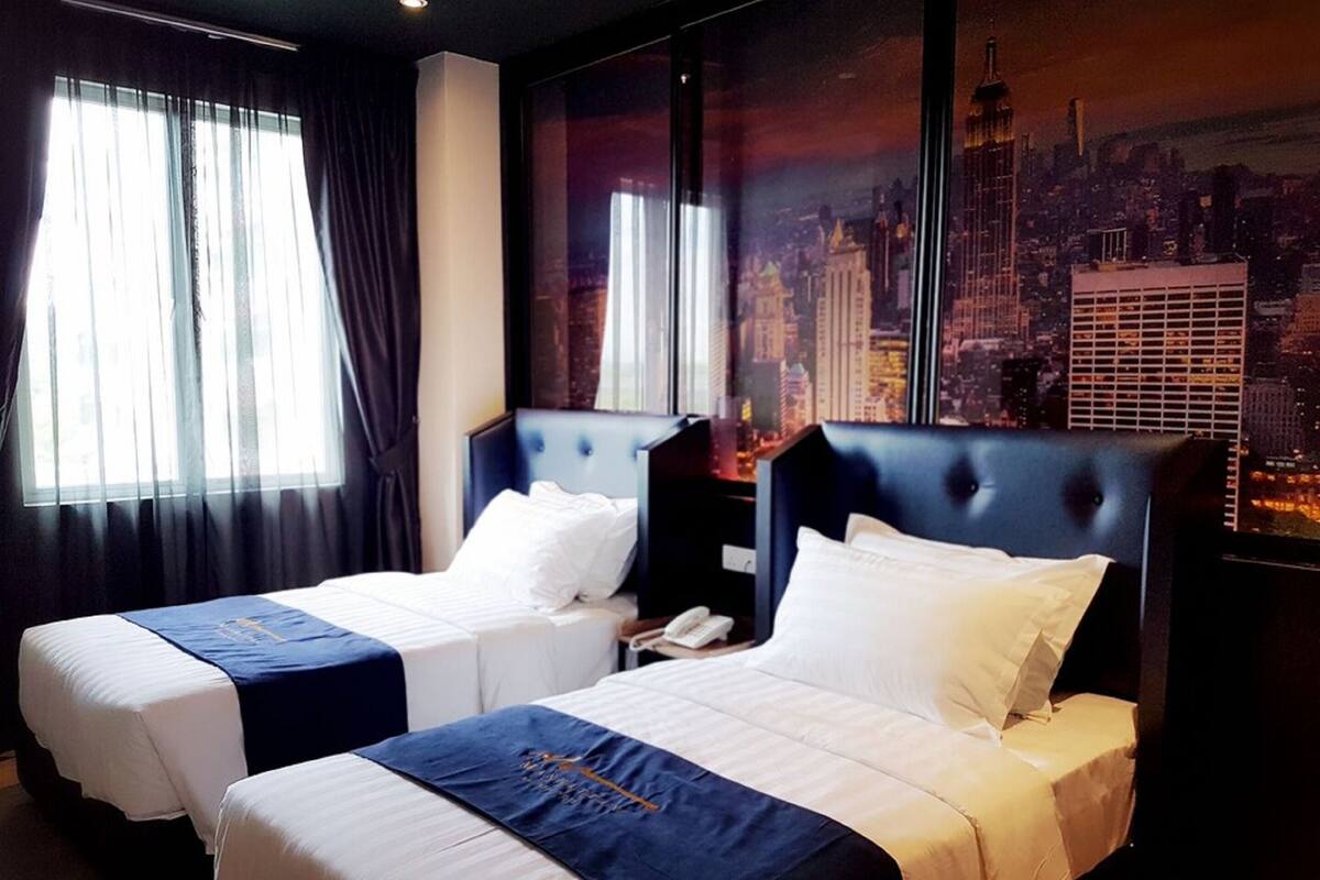 Une chambre d’hôtel avec deux lits, chacun doté d’une tête de lit bleue et de draps blancs. La chambre est pourvue de grandes fenêtres avec des rideaux légers, qui laissent entrer la lumière naturelle. Une fresque représentant un paysage urbain est visible sur le mur derrière les lits.