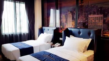 Habitación de hotel con dos camas, cada una con cabecero azul y ropa de cama blanca. La habitación tiene ventanas grandes con cortinas translúcidas que dejan entrar luz natural. Se ve un mural con un paisaje urbano en la pared detrás de las camas.