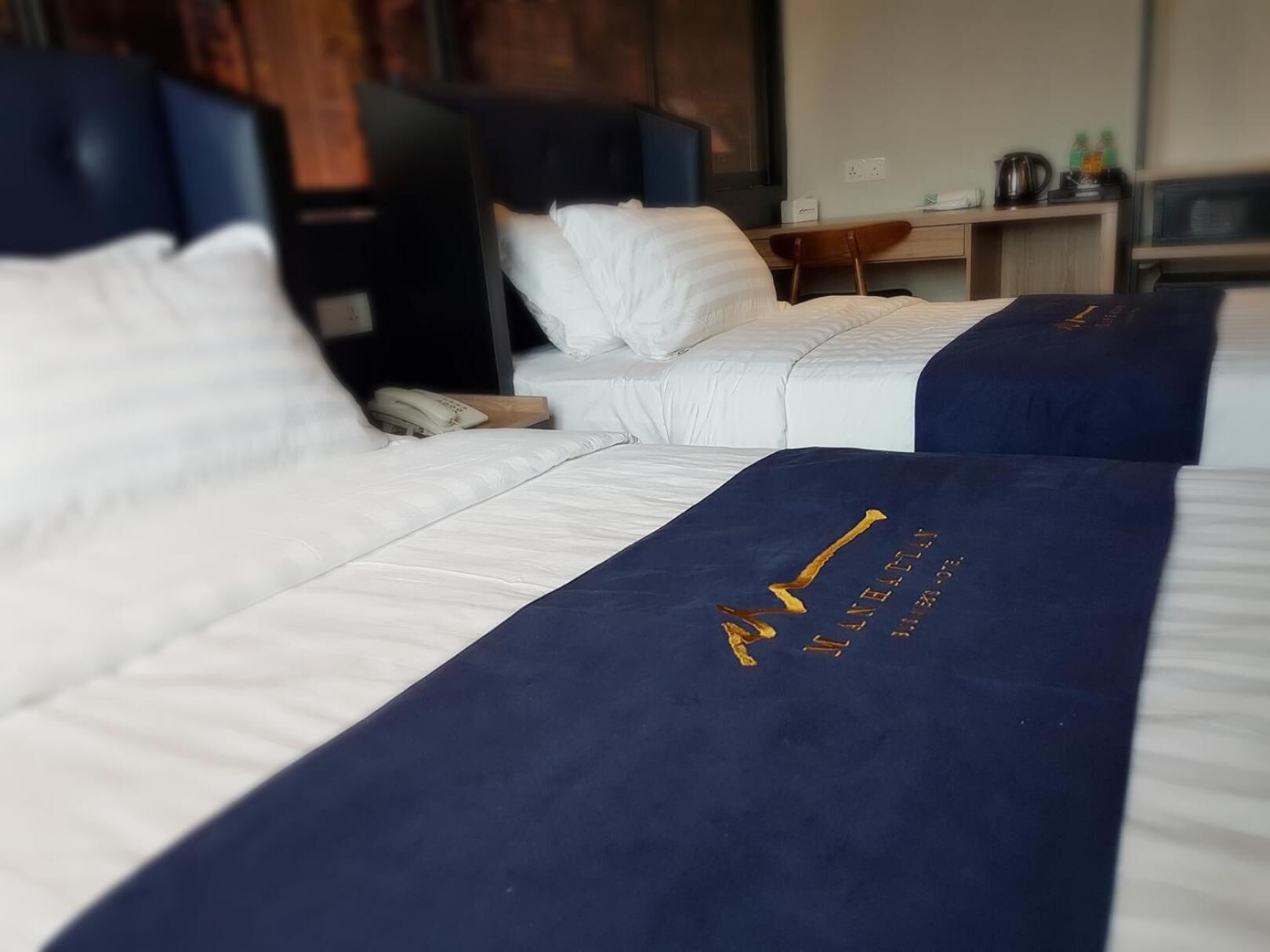 Ein Hotelzimmer mit zwei Betten, jeweils mit weißer Bettwäsche und einer dunkelblauen Tagesdecke. Auf einem kleinen Tisch stehen ein Wasserkocher und ein Telefon.