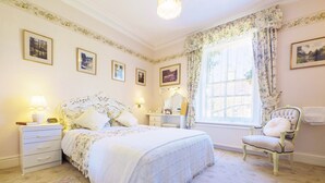 A classic bedroom with a large bed, a nightstand, a dresser, a chair, and a window with curtains.