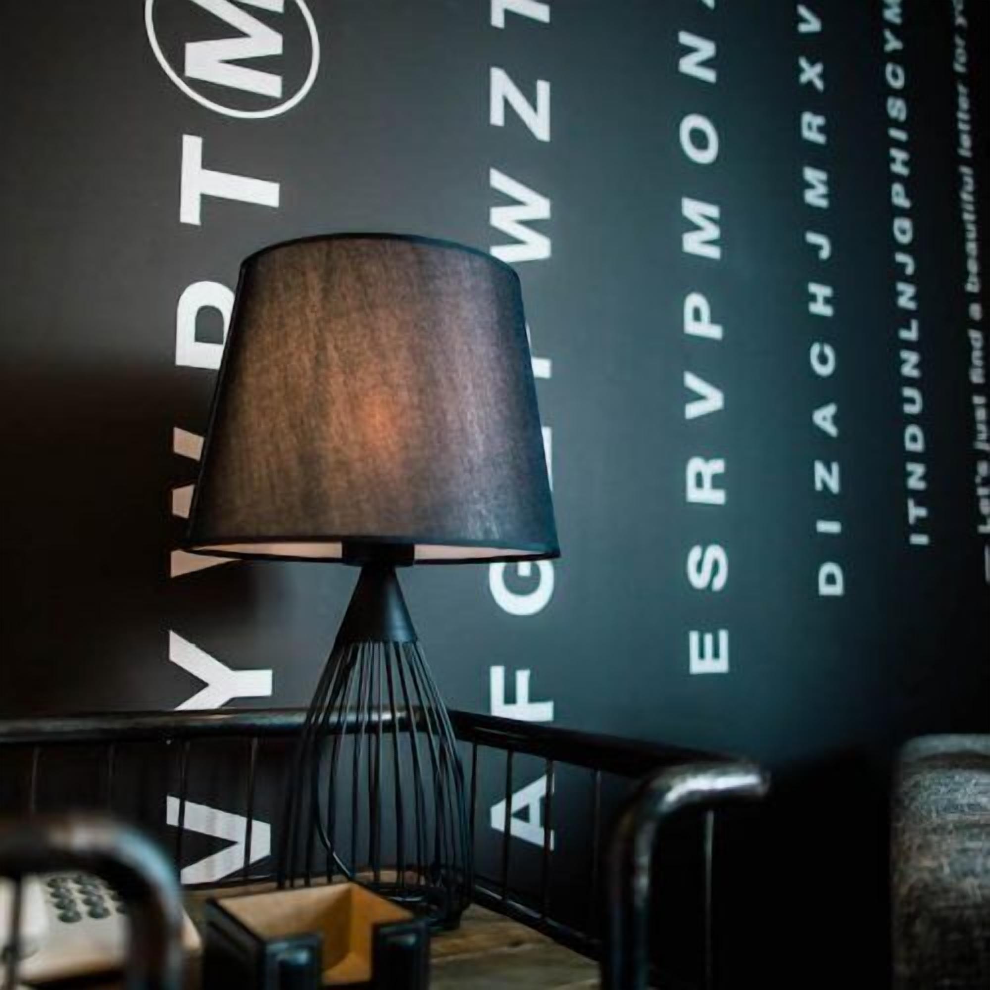 Una lámpara moderna con una pantalla oscura, un soporte de metal negro y un logotipo visible en la pared de fondo.