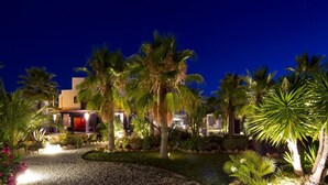 A nighttime view of a resort with palm trees, a well-lit pathway, and a building with a red-lit entrance. - Hotel Boutique Ses Pitreras (Sant Josep de sa Talaia)