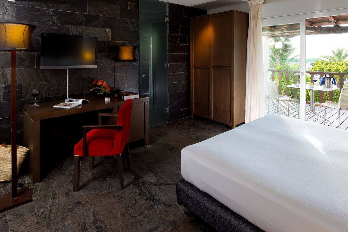 A hotel room with a bed, desk, chair, TV, and a balcony with outdoor seating.