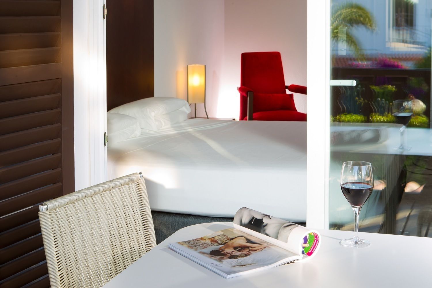 Una habitación de hotel con una cama, una silla roja, una mesa con una copa de vino y una revista.