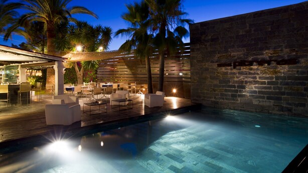 A poolside area with lounge chairs and tables, a stone wall, and palm trees. - Hotel Boutique Ses Pitreras (Sant Josep de sa Talaia)