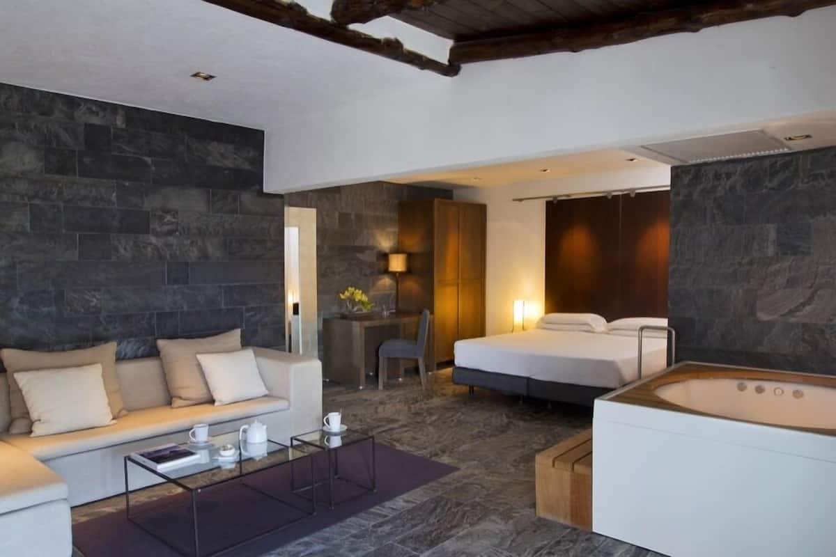 A modern hotel room with a stone wall, a sofa, a bed, a desk with a lamp, and a bathtub.