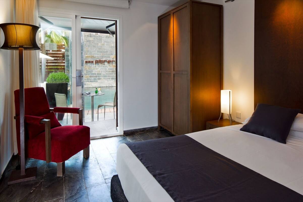 A hotel room with a bed, a red chair, a floor lamp, and a view through a glass door to an outdoor area with a table and chairs.