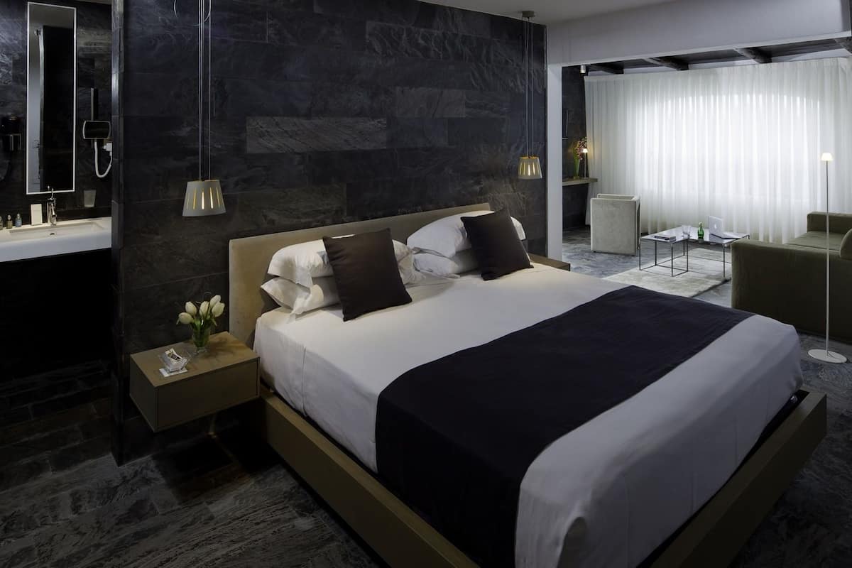 A modern hotel room with a large bed, a bedside table with a lamp, a mirror, and a bathroom with a shower and a toilet.
