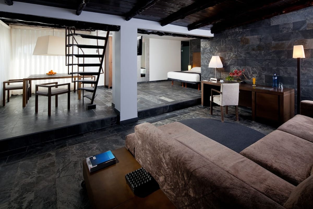 A modern hotel room with a loft-style bed, a desk with a lamp, a chair, and a laptop on a small table.