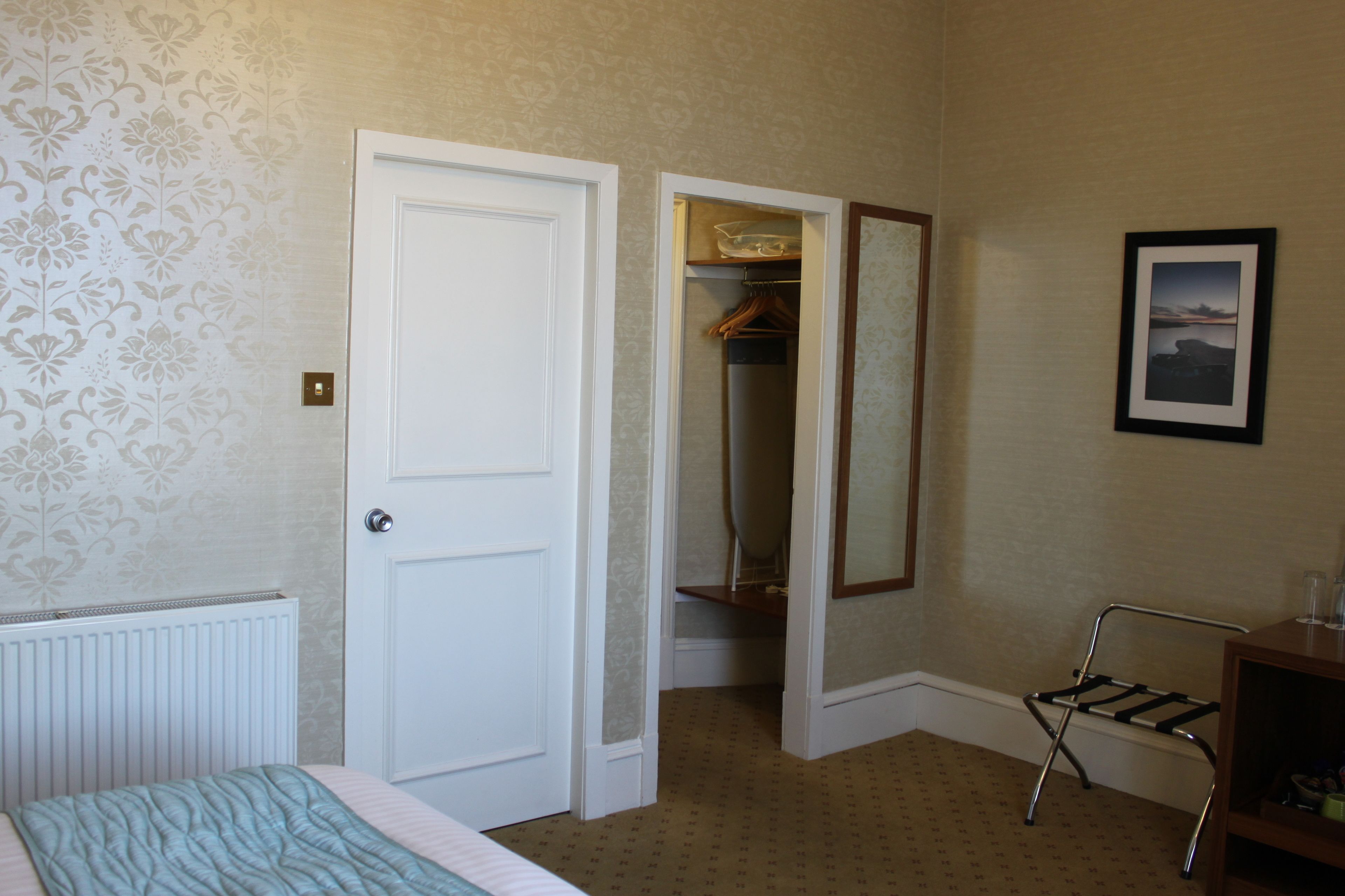 A hotel room with a bed, a radiator, a wardrobe, a mirror, and a shelf with items.