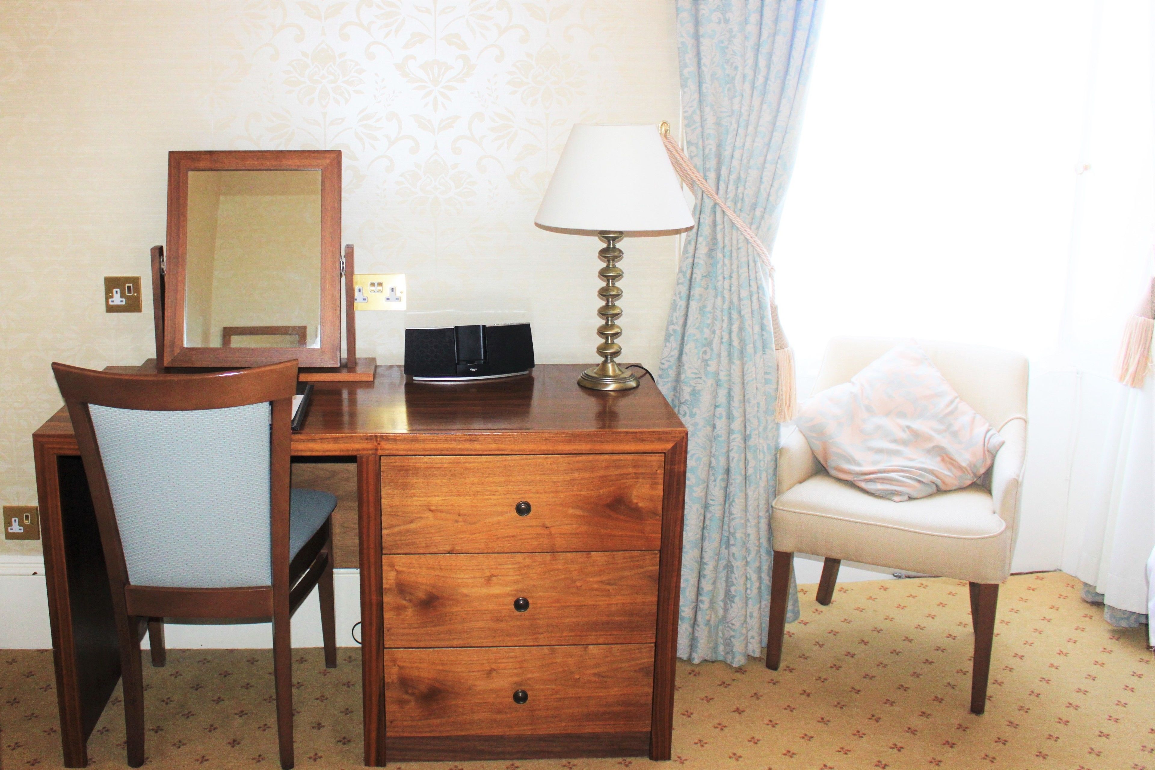 A wooden desk with a mirror, a lamp, and a chair in a room with patterned wallpaper and a carpet.