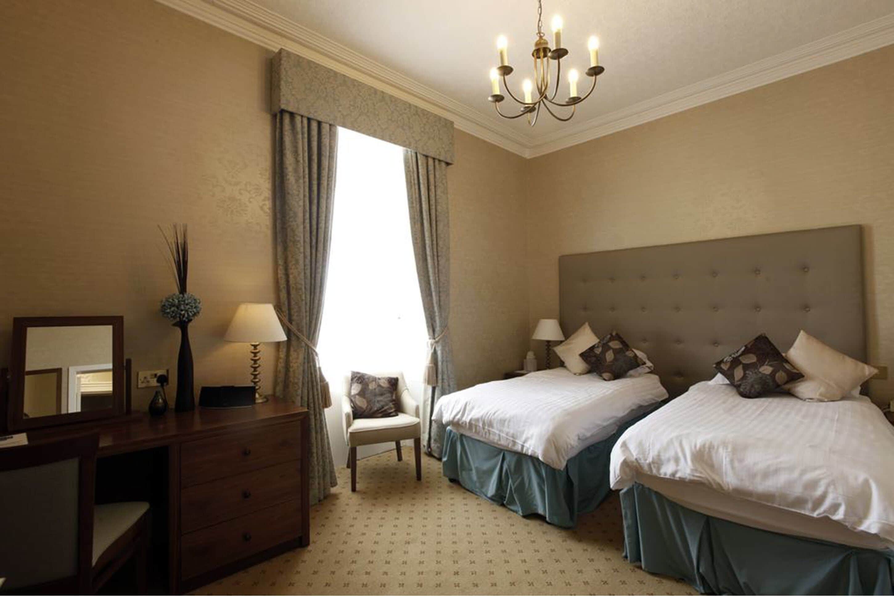 A hotel room with two beds, a desk, a chair, a chandelier, and a large window with curtains.