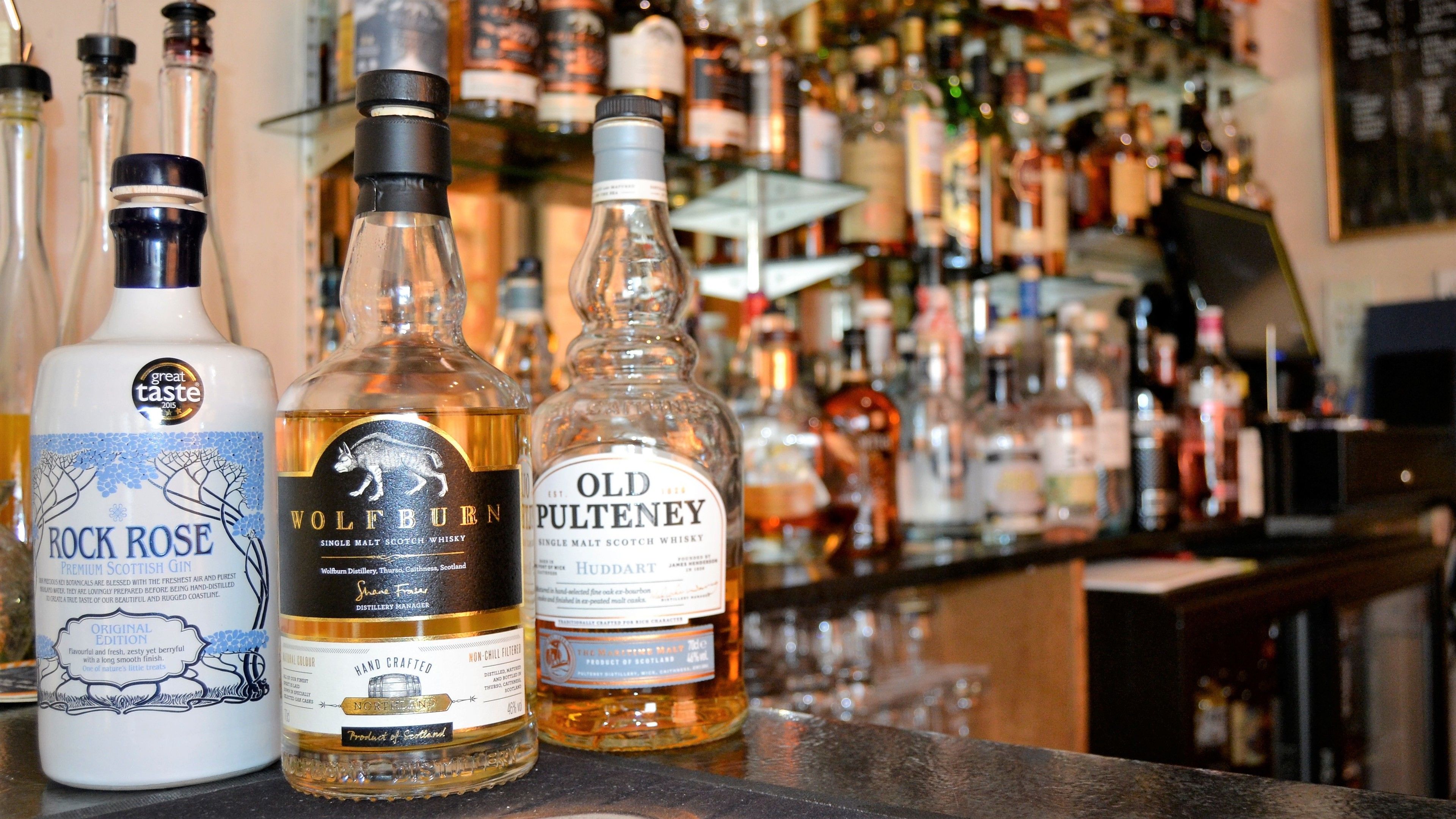Three bottles of whisky on a bar counter with a blurred background of various bottles.