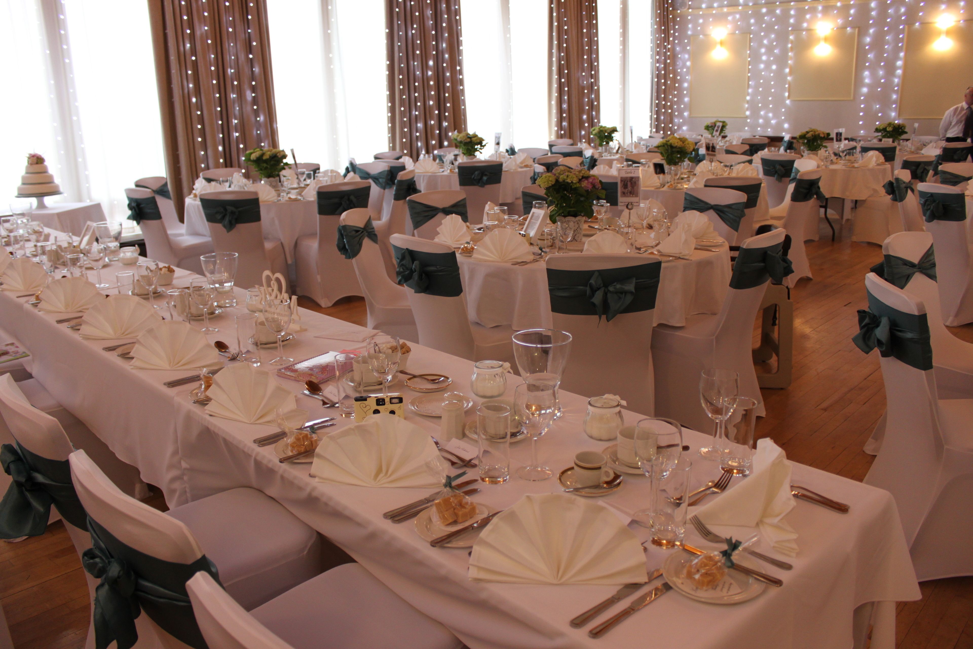 Un salón de banquetes con mesas dispuestas para un evento formal, destacando manteles blancos, cintas verdes y centros de mesa florales.
