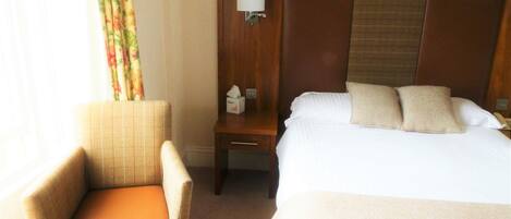 A hotel room with a bed, a chair, a nightstand with a lamp, and a window with curtains.