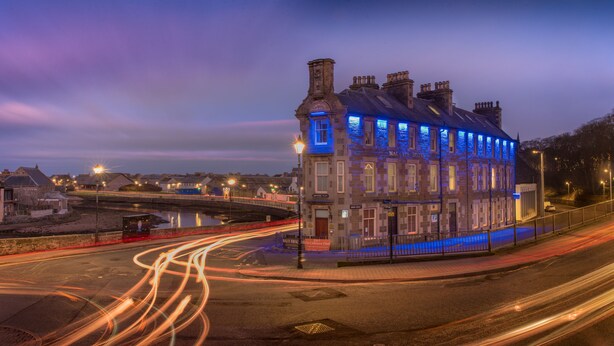 A historic building with a prominent tower and multiple chimneys, located at a street corner with a view of a river. - Mackays Hotel Wick (Wick)