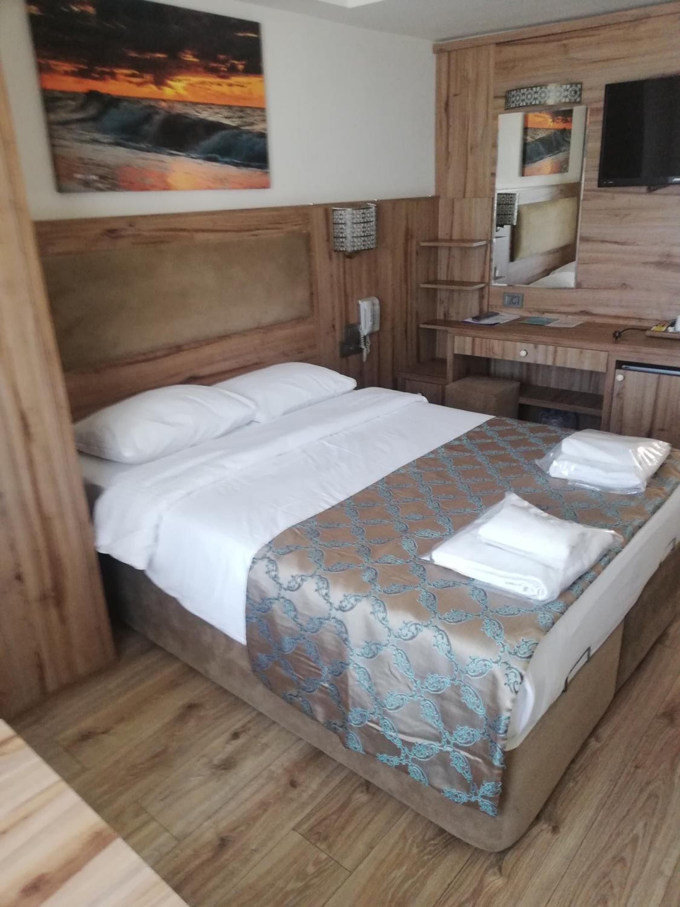 Habitación de hotel con cabecera de madera, cama con ropa blanca, colcha estampada, cómoda de madera, espejo y un cuadro en la pared.