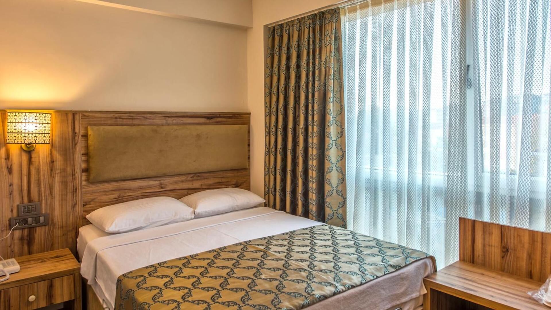 Una habitación de hotel con cabecera de madera, una cama con colcha estampada, una mesita de noche con lámpara y una ventana con cortinas translúcidas.