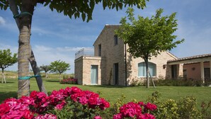A two-story stone building with a balcony, surrounded by well-maintained grass and flowering plants. - Officina del Sole (Montegiorgio)