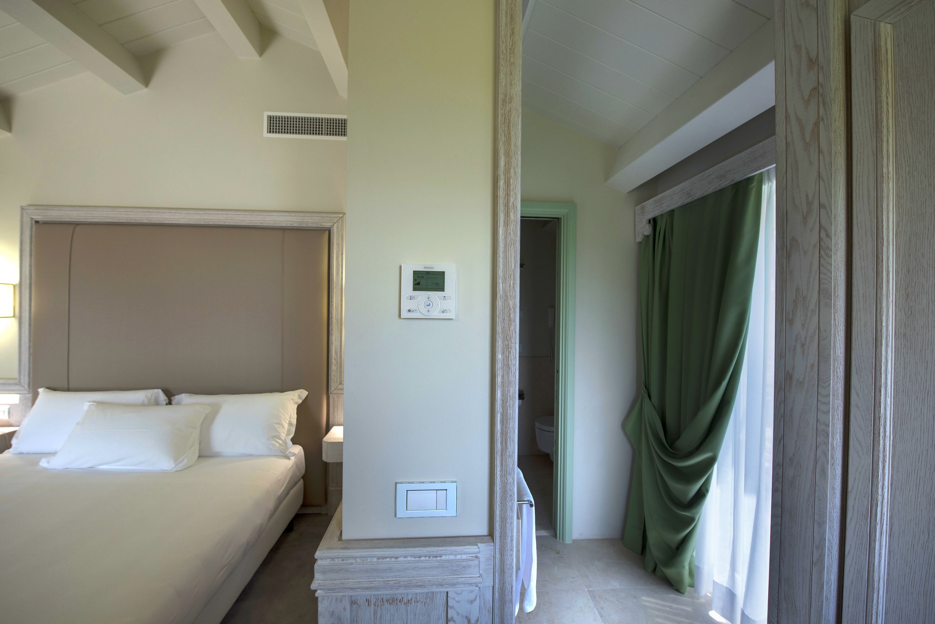 A hotel room with a bed, a thermostat, and a view of the bathroom.