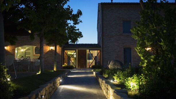 A pathway leading to a building with a glass door, surrounded by stone walls and greenery. - Officina del Sole (Montegiorgio)