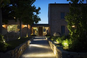 A pathway leading to a building with a glass door, surrounded by stone walls and greenery. - Officina del Sole (Montegiorgio)