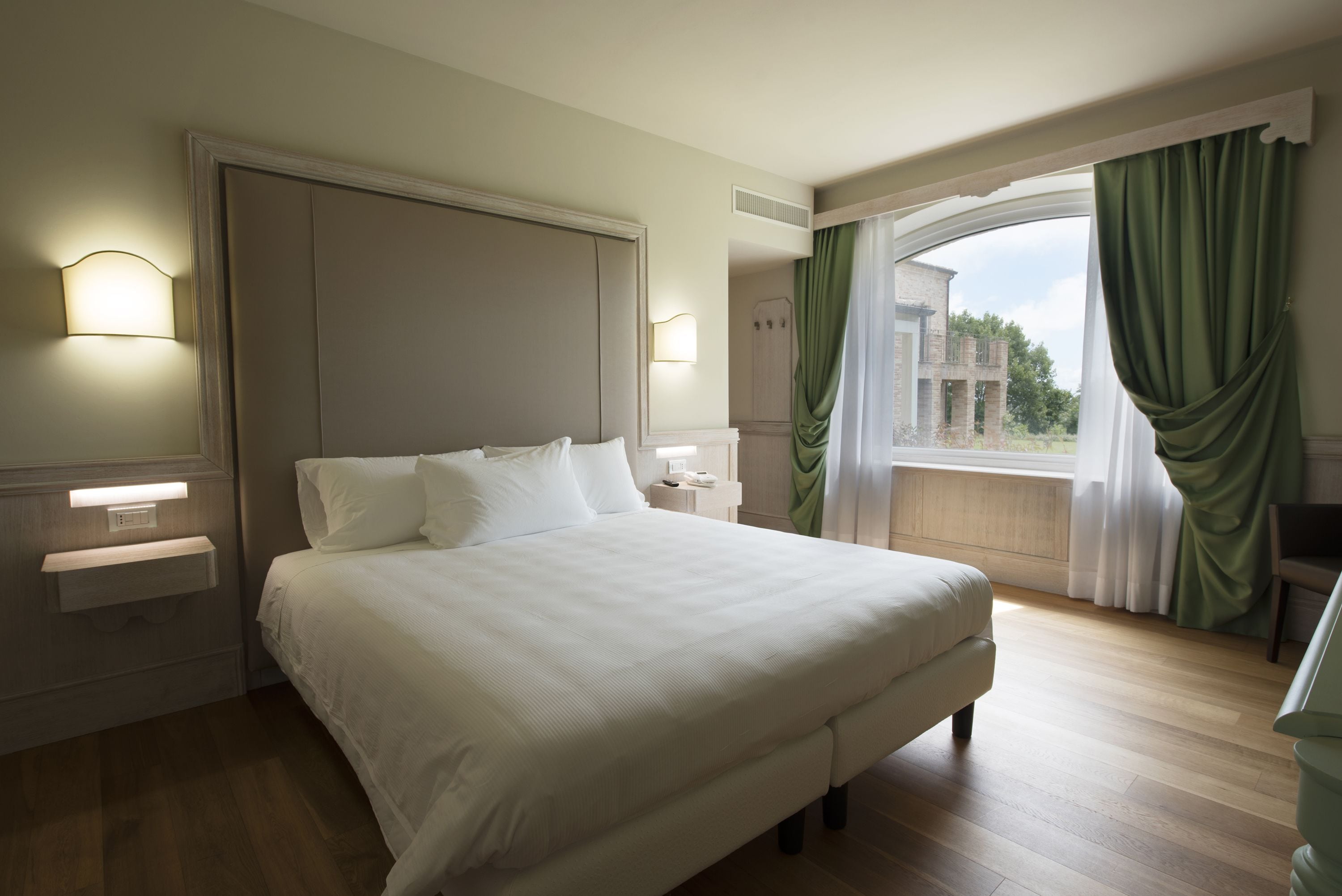 A hotel room with a large bed, bedside lamps, a nightstand, and a view of a building through a window with green curtains.