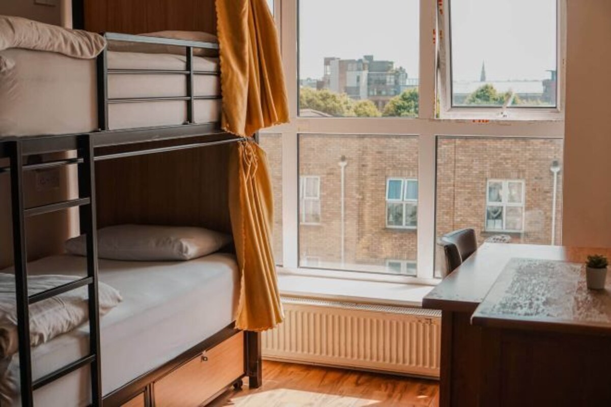 Een studentenkamer met stapelbedden, een bureau en uitzicht op de stad door grote ramen.