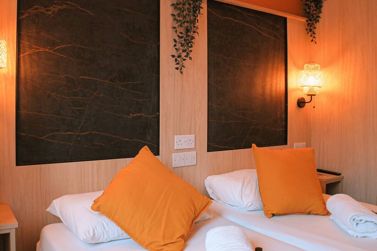 A hotel room with two beds, each with white bedding and orange pillows. There are wall-mounted light fixtures and a plant hanging above the beds.