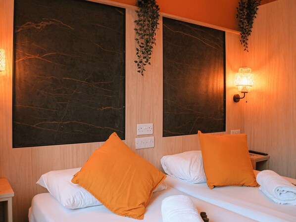 Habitación de hotel con dos camas, cada una con ropa blanca y almohadas naranjas. Hay luces fijadas a la pared y una planta colgando sobre las camas.