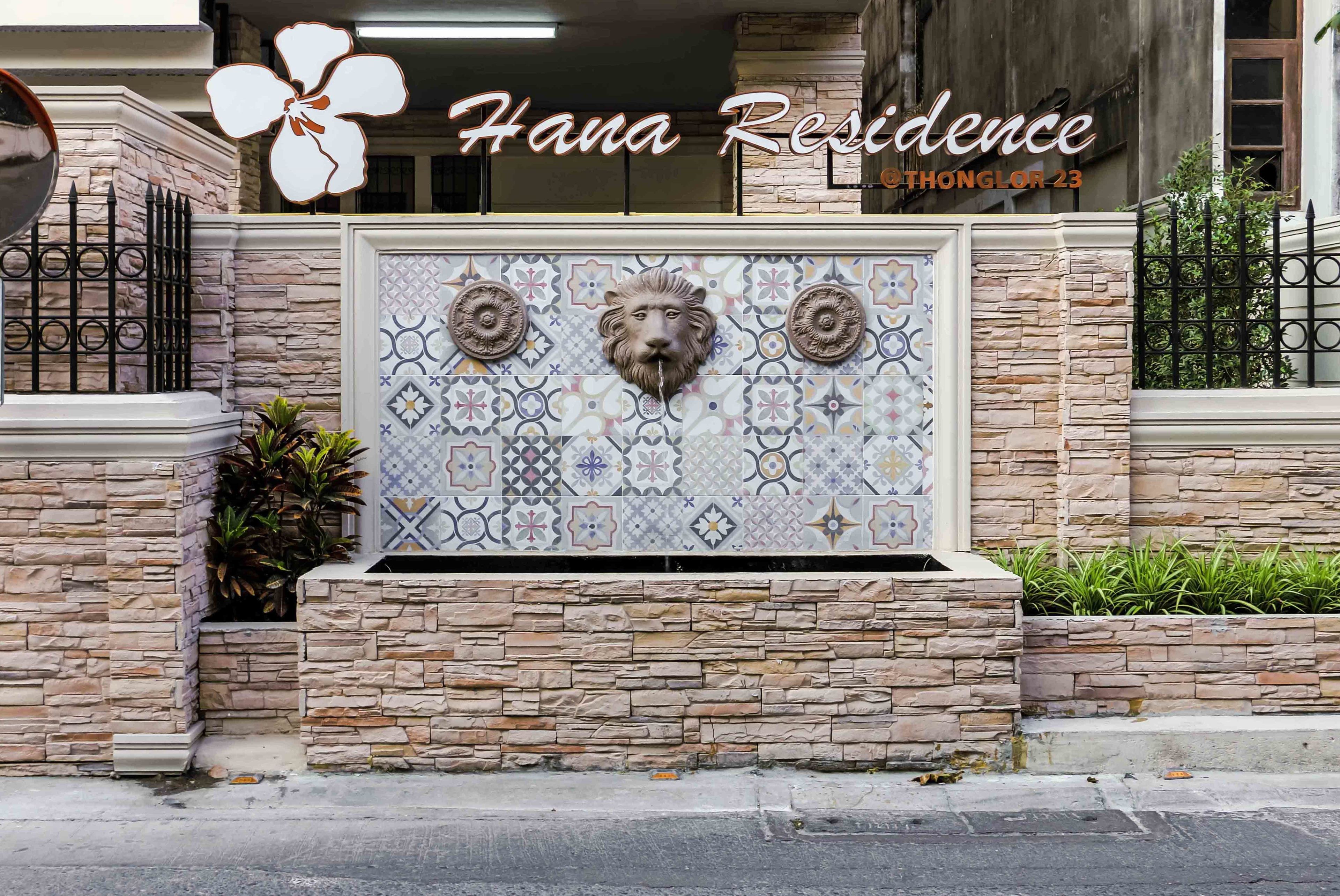 A building facade with a stone wall, a decorative tiled panel with lion heads, and a sign reading "Hana Residence THONGLOOR 23".