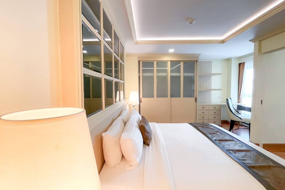 A bedroom with a large bed, a nightstand, a desk, and a chair. There is a window with blinds and a ceiling light.