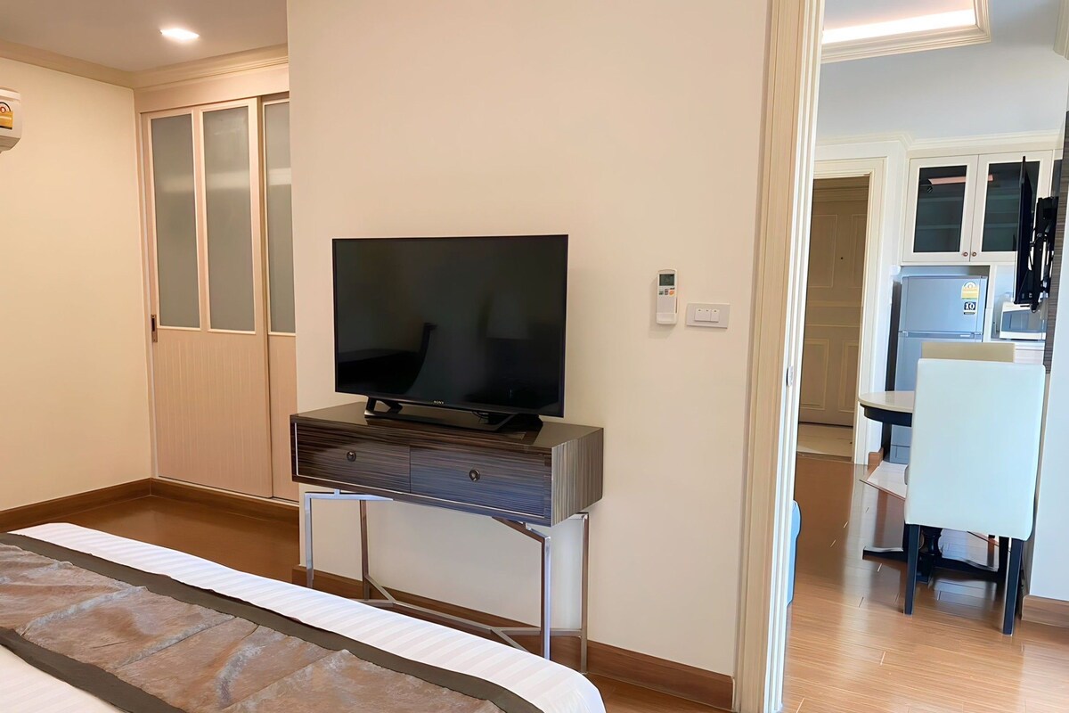 A hotel room with a bed, a TV on a stand, a desk with a chair, and a kitchenette.