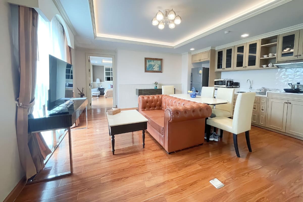 A modern living room with a leather sofa, a coffee table, and a TV. There is a dining area with a table and chairs, and a kitchen with cabinets and appliances.