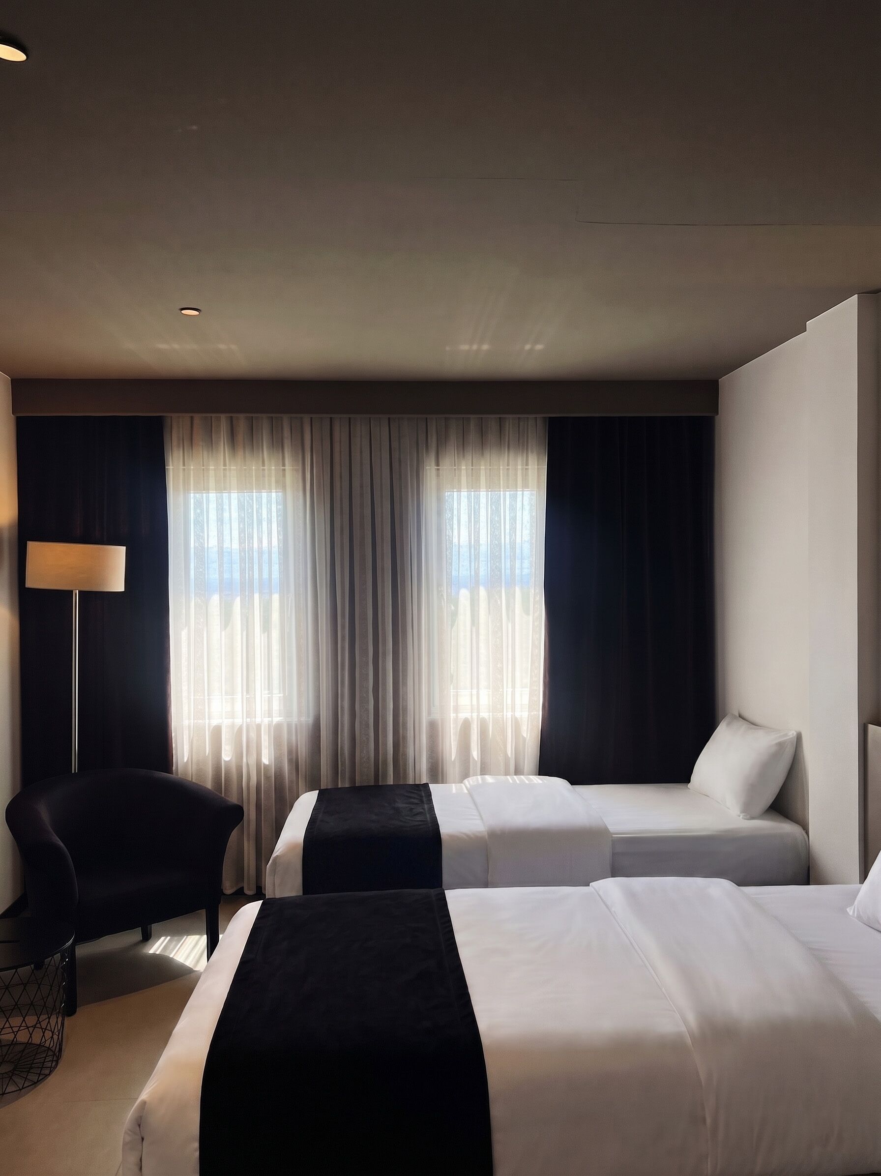 Habitación de hotel con dos camas, una silla, una lámpara y un ventanal grande con cortinas.