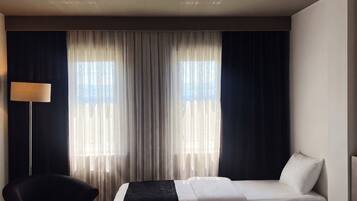 Habitación de hotel con dos camas, una silla, una lámpara y un ventanal grande con cortinas.