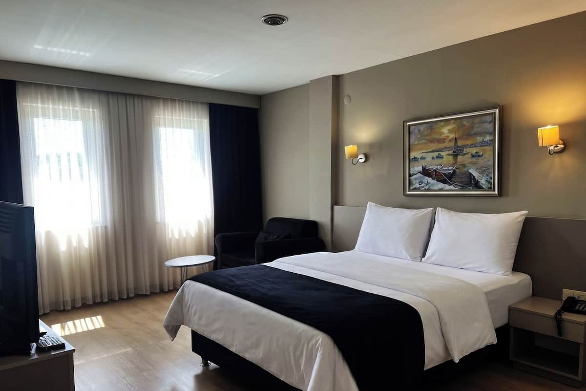 Une chambre d’hôtel avec un grand lit, un bureau avec un ordinateur, une chaise, un tableau au mur et une fenêtre avec des rideaux.