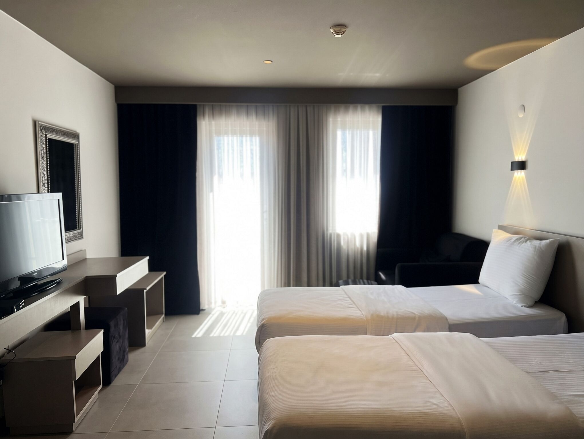 Een hotelkamer met twee bedden, een bureau, een televisie en een groot raam met gordijnen.