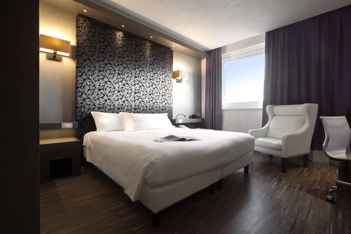 Une chambre d’hôtel moderne avec un grand lit, un fauteuil blanc et une vue sur le ciel par la fenêtre.
