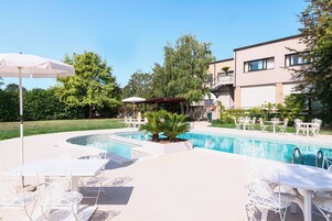 A swimming pool with white outdoor furniture, a covered seating area, and a building in the background.