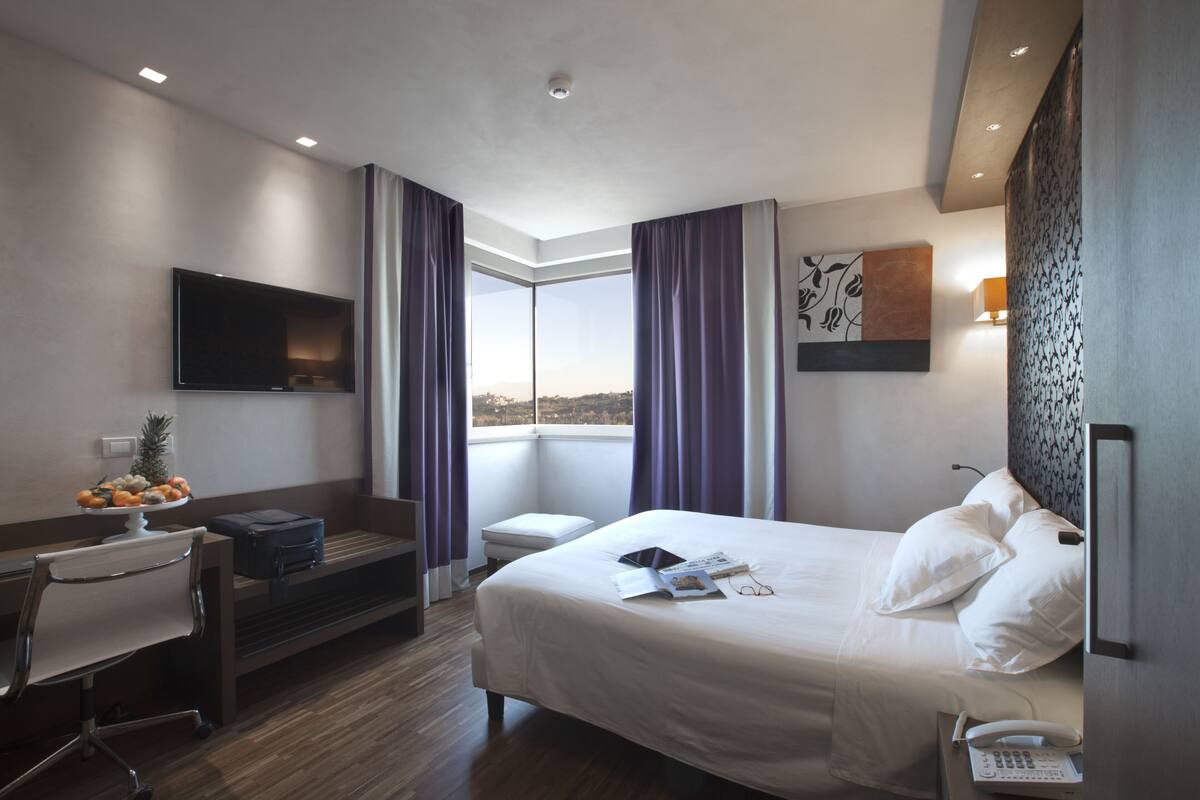 Une chambre d’hôtel avec un lit, un bureau, une chaise, une télévision, une fenêtre avec des rideaux et une vue sur la ville.