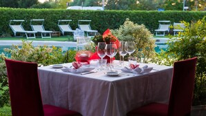 Mesa posta para dois, com toalhas brancas, cadeiras vermelhas e vista para uma piscina e vegetação ao fundo.