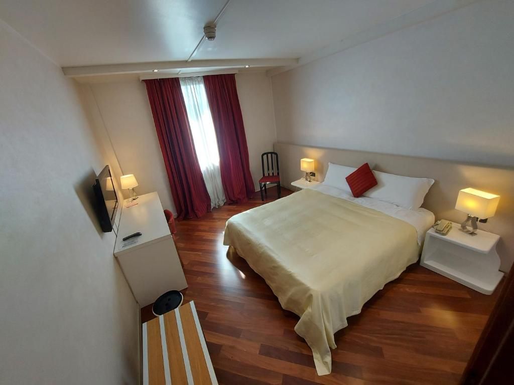 Una habitación de hotel con cama, mesitas de noche, televisor, escritorio y ventana con cortinas.