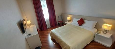 Una habitación de hotel con cama, mesitas de noche, televisor, escritorio y ventana con cortinas.
